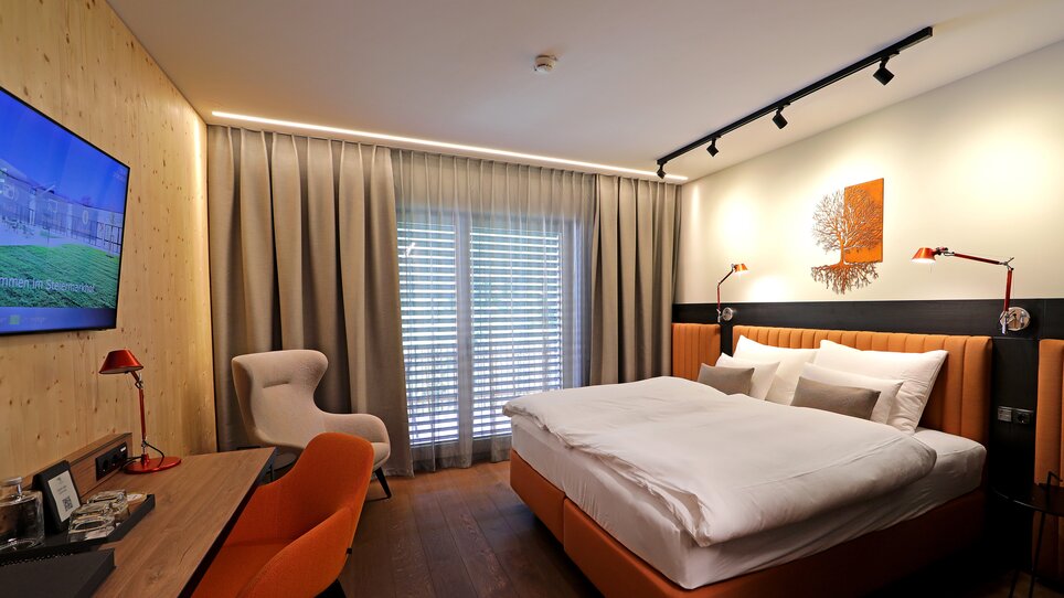 Modernes Doppelzimmer mit Bett, Schreibtisch, Fernseher und großer Fensterfront. Der Raum ist in warmen Beige- und Orangetönen gestaltet. | © Hotel Steiermarkhof