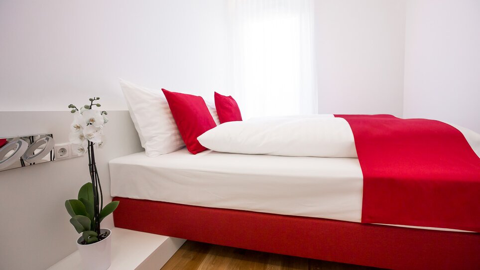 Stanza luminosa con letto matrimoniale e cuscini rossi. | © sHome Hotel