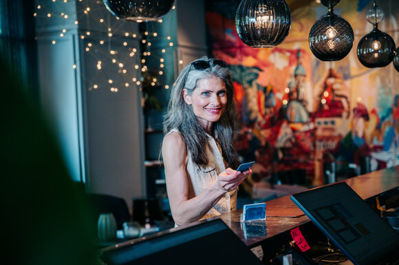 Eine Frau mit langen Haaren hält ein Handy in der Hand und steht an der Hotel-Rezeption. | © Graz Tourismus - Mias Photoart