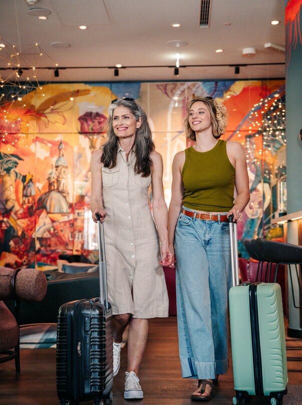 Zwei Frauen mit Koffern in einer bunten Hotelumgebung. | © Graz Tourismus - Mias Photoart